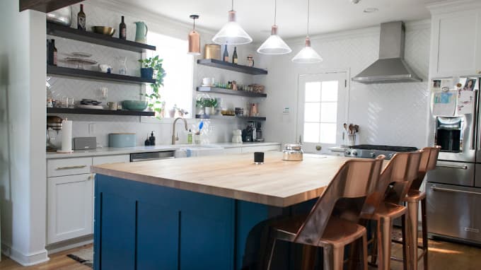 This is an image from JT Paint & Design, located in Oklahoma. The photo shows a stylish kitchen with a large wooden island, blue cabinetry, white backsplash, open shelving with décor, and pendant lighting over bar seating.