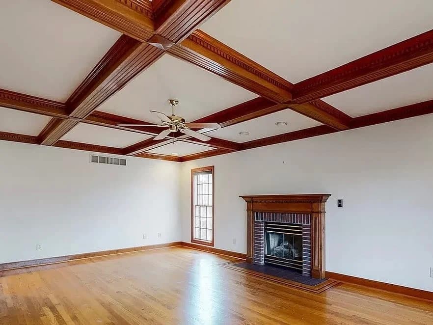 This is an image from Alarcon Pro Painting & Design, located in North Carolina. The image shows a bright living room with a coffered wooden ceiling, a ceiling fan, hardwood floors, and a fireplace with a wooden mantel.