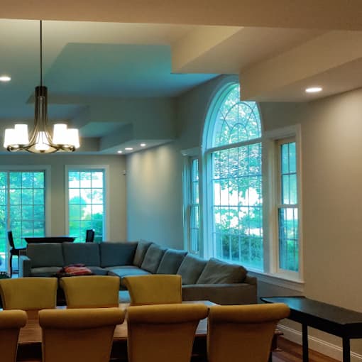 This is an image from Big Expert Painting LLC, located in New York. The interior photo shows a living and dining area with freshly painted light-colored walls, recessed ceiling lighting, large arched windows, and a sectional sofa arranged near a dining table.