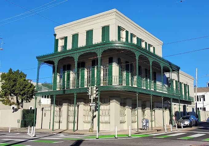This is an image from Pelican Paint Group, located in Louisiana. The photo shows a historic three-story corner building with light beige walls and dark green shutters, featuring ornate wrought-iron balconies typical of classic New Orleans architecture.