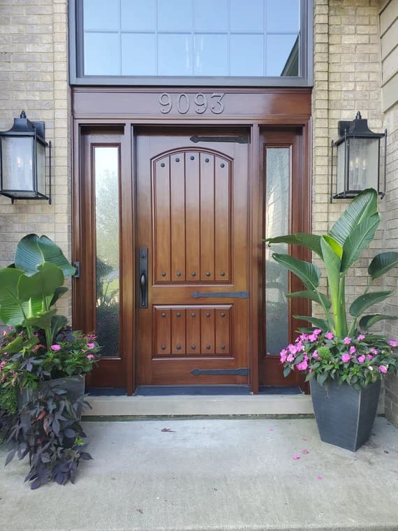 This is an image from MacFarland Painting - Ann Arbor company, located in Ann Arbor, MI The image is a refinished wooden front door with decorative iron accents and side glass panels, completed by MacFarland Painting, surrounded by potted plants with pink flowers.