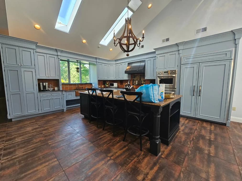 This is an image from JRT Pro Painters, located in New Jersey. The image shows a completed kitchen painting project with custom gray cabinetry, a black island, wood flooring, and modern lighting.