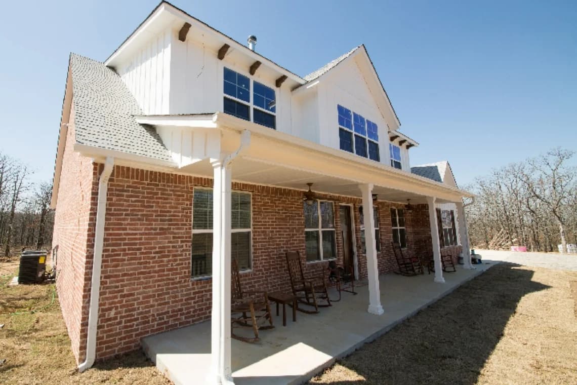 White and red brick farmhouse painted by Remarkable Painting, with crisp white siding on the upper level, large front porch, and wooden rocking chairs creating a welcoming appearance.
