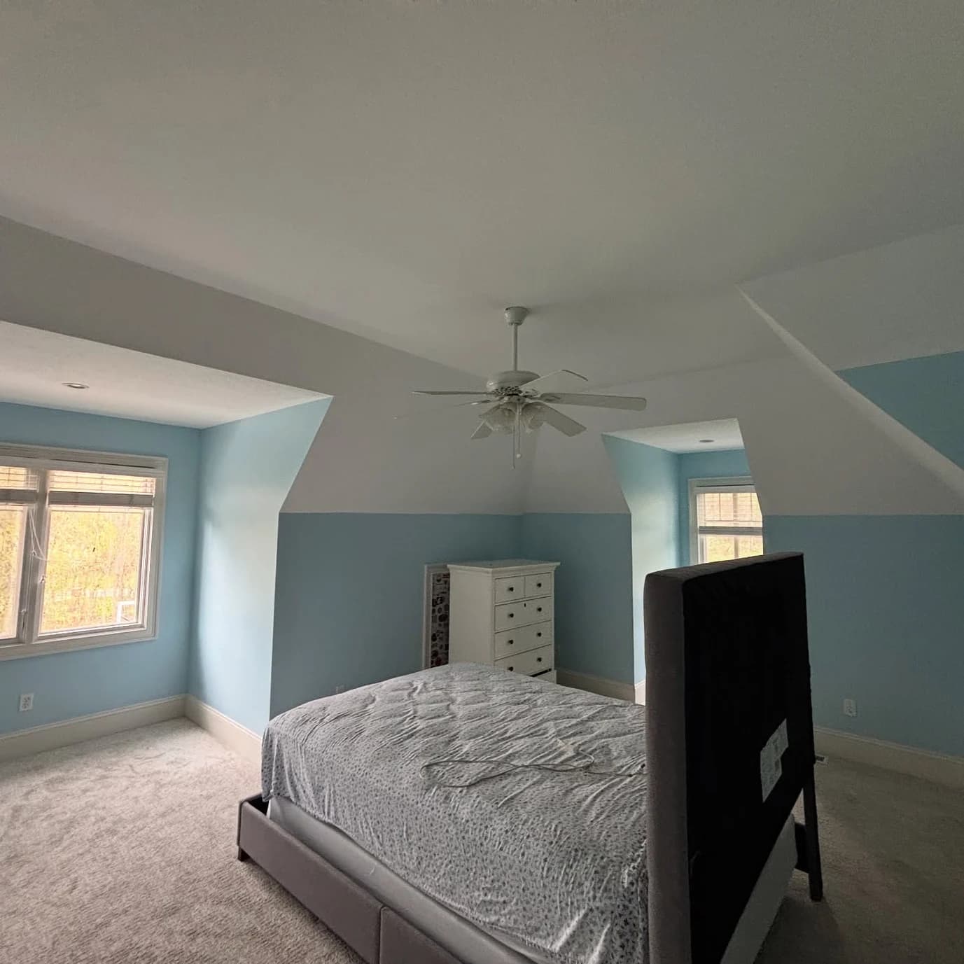 This is an image from 216 Painting, located in Ohio. The image shows a bedroom interior with soft blue walls, white trim, vaulted ceilings, a ceiling fan, and natural light coming through multiple windows.