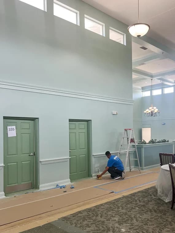 This is an image from Halls Quality Painting company, located in Fernandina Beach, FL. The image is a professional interior painter applying final touch-ups on light green doors and walls in a high-ceiling commercial space with decorative molding.