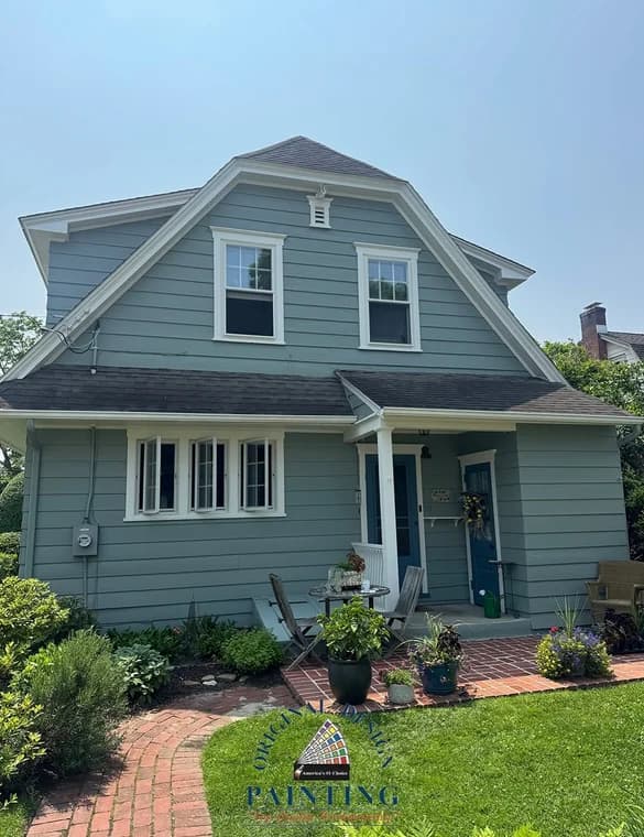 This is an image from Original Design Painting, showcasing a light blue two-story home with white trim. The house has a steep roofline and a welcoming brick walkway leading to the entrance.