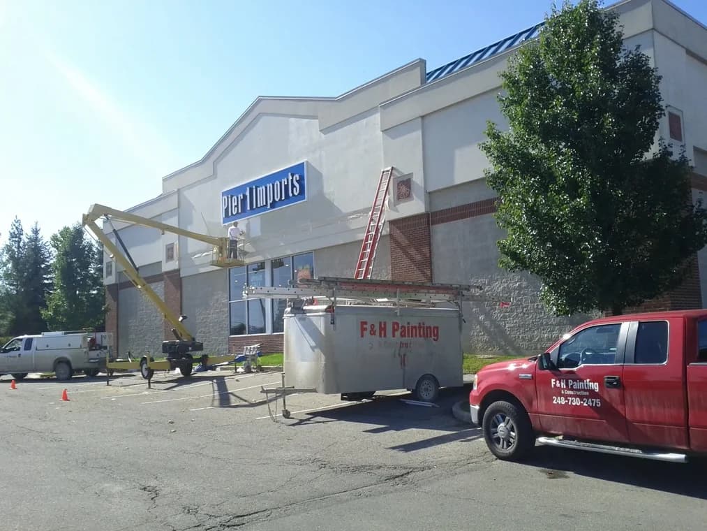 This is an image from The Exterior Painting Experts - F&H Painting company, located in Oxford, MI The image is a F&H Painting crew performing exterior commercial painting at Pier 1 Imports, featuring red company truck and trailer with phone number 248-730-2475, and a worker on a hydraulic lift.