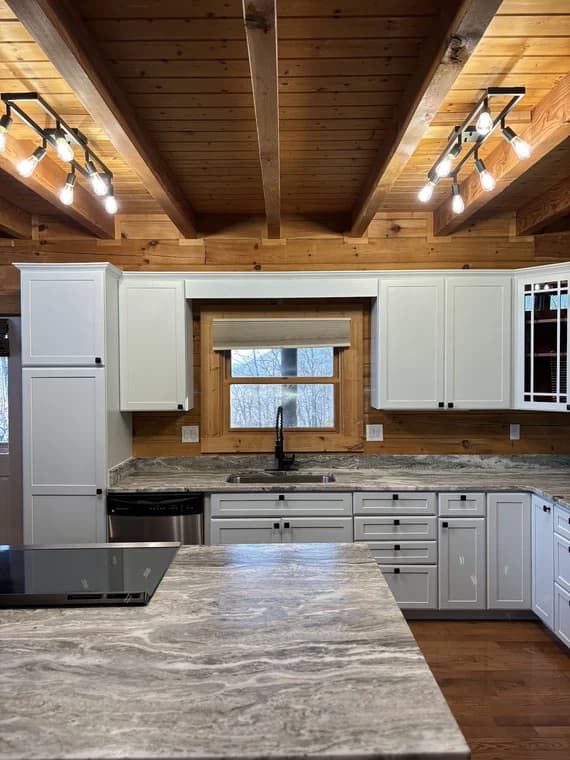 Cabinet painting project by Passion Painters in Swannanoa, NC. The image shows white kitchen cabinets, wood walls and ceiling beams, and gray stone counters.