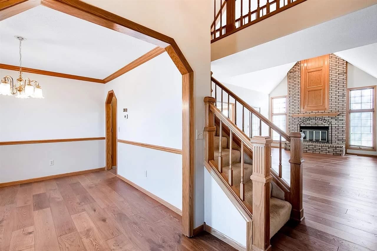 This is an image from Northwind Painting LLC. The image shows an interior repaint featuring fresh white walls and trim throughout the entry, stairway, and living room, highlighting the natural woodwork and brick fireplace.