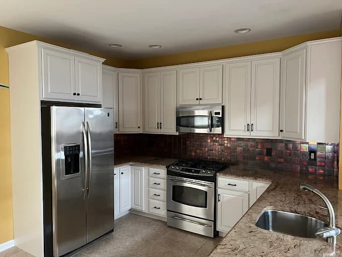This is an image from Willow Tree Painting & Home Improvement LLC, located in New York. The photo shows a modern kitchen with freshly painted white cabinets, stainless steel appliances, and a dark textured backsplash.