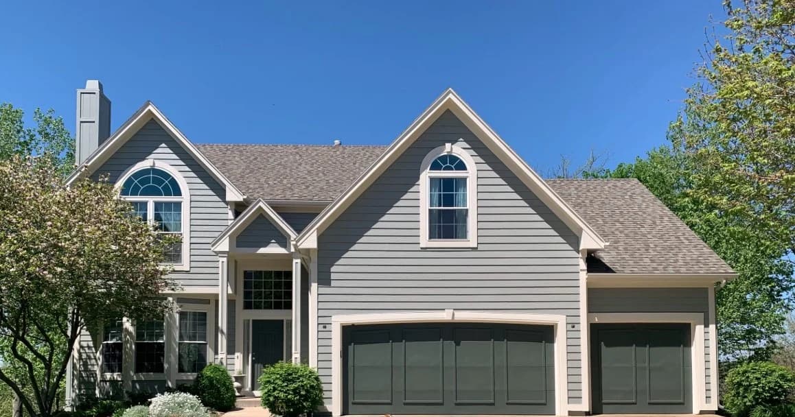 This is an image from CertaPro Painters of Shawnee Mission, KS. The image is a two-story light gray home in Shawnee Mission KS with fresh exterior paint and dark garage doors.