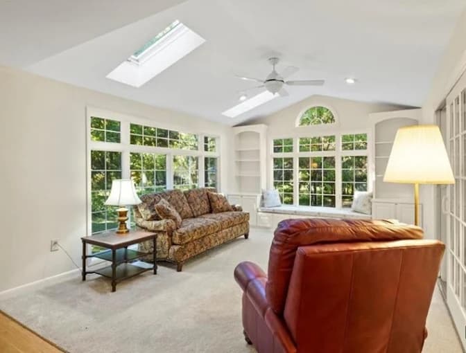 This is an image from Monarch Painting, located in New York. The photo shows a bright living room with large windows, skylights, neutral-colored walls, and furnished seating including a patterned sofa and a red leather chair.