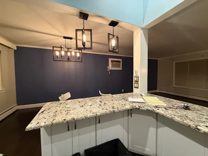 This is an image from Paul’s Painting & Powerwashing, located in New Jersey. The image shows a renovated kitchen area with painted cabinetry, a granite countertop, and modern pendant lighting above a breakfast bar.