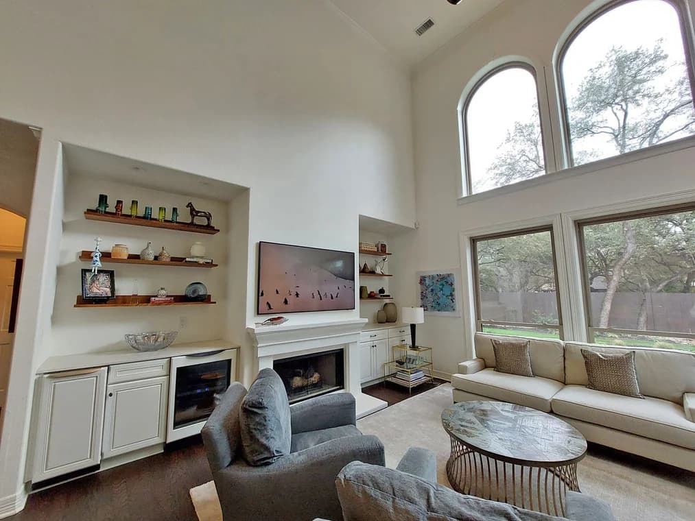 "This is an image from Paramo Painting, located in Texas. The living space includes built-in shelving, a fireplace, large windows facing the yard, and a seating area with a sofa and armchairs."