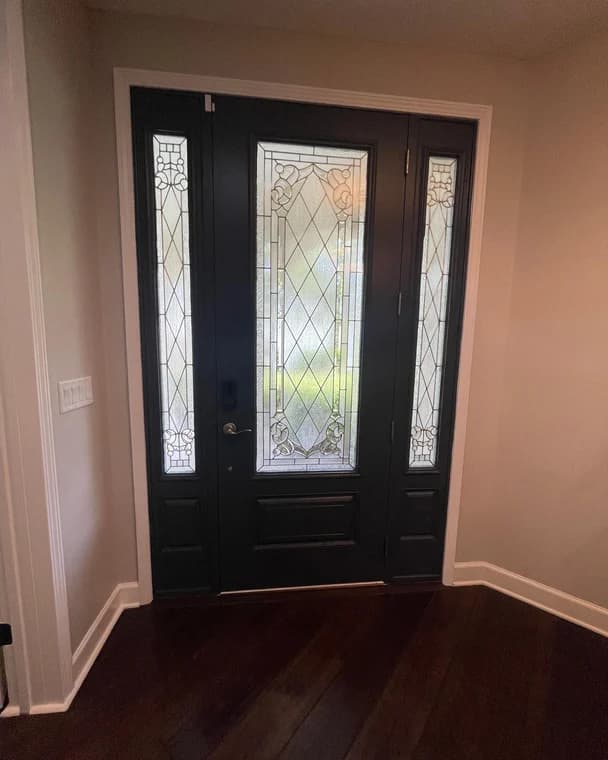 This is an image from Bright Side Painting Of Jax company, located in Jacksonville Beach, FL. The image is a freshly painted dark wood front door with decorative frosted glass panels, enhancing the entryway of a modern home interior with rich hardwood floors