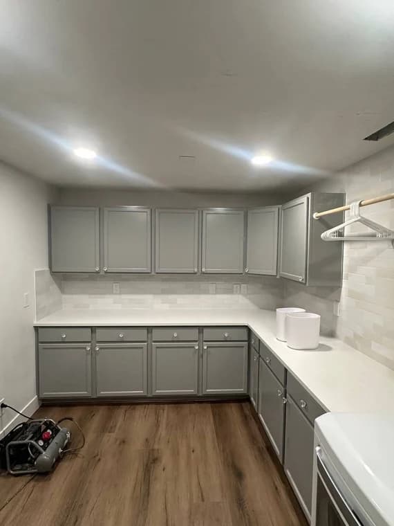 This is an image from Montenegro's Painting & Construction LLC company, located in Meridian, ID The image is a modern laundry room with freshly painted gray cabinets, white quartz countertops, subway tile backsplash, wood-look vinyl flooring, and overhead LED lighting, refinishing by Montenegro's Painting & Construction, LLC