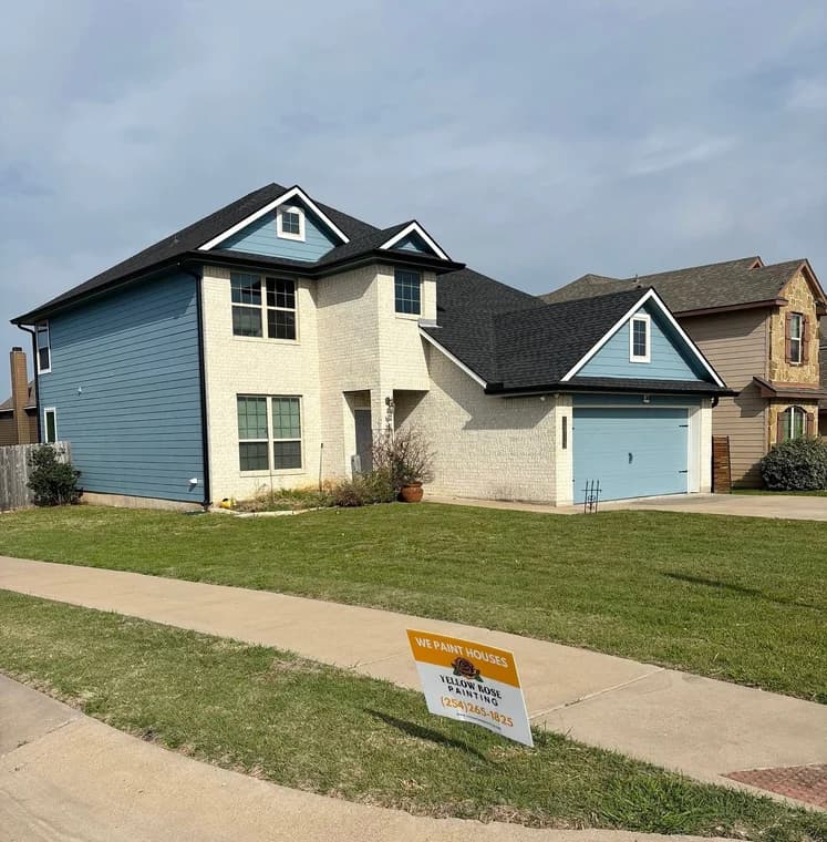 This is an image from Yellow Rose Painting, located in Texas. The home exterior displays blue siding, light brickwork, and a matching blue garage door with a yard sign advertising the company's painting services.