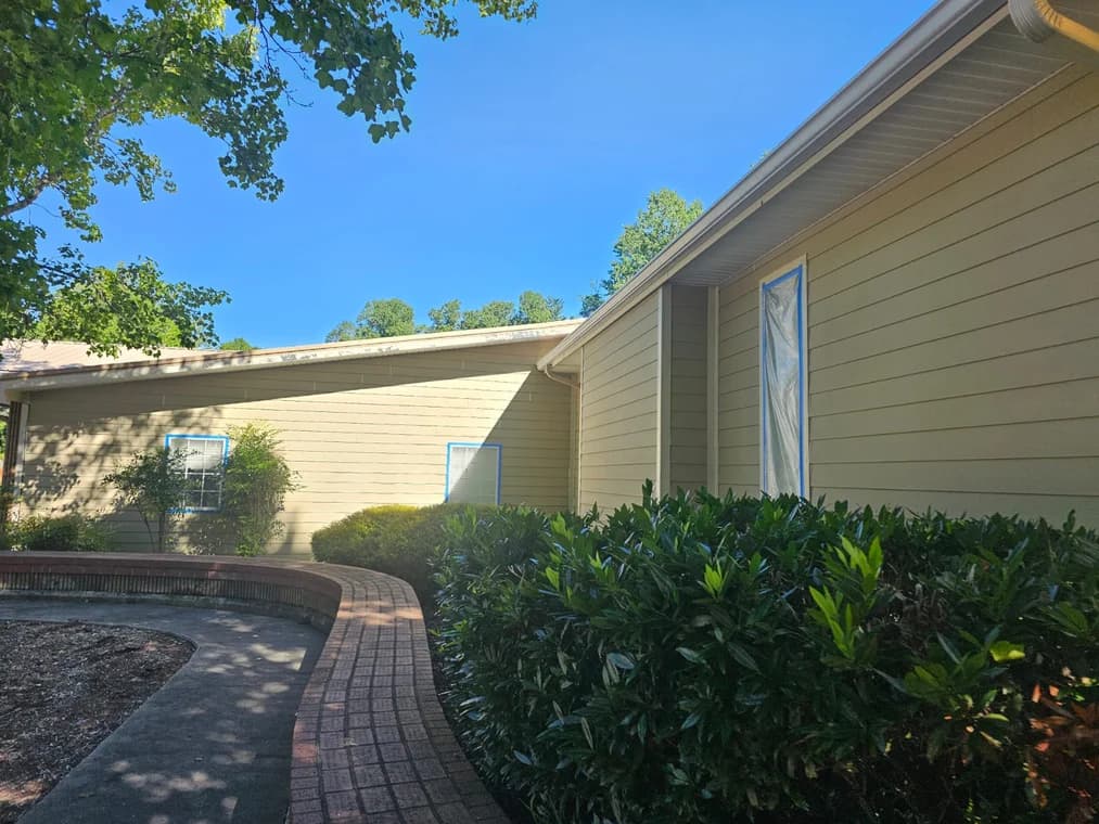 Exterior painting project by Custom Painting of Asheville inc in Asheville, NC. The image shows tan siding, white trim, taped windows, shrubs, and a walkway.