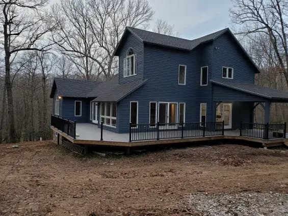 This is an image from Sam Professional Painting LLC, located in Maryland. The exterior home photo shows a blue two-story house with a wraparound deck and painted trim surrounded by woodland.