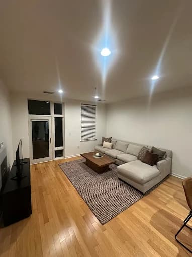 This is an image from By Hand Painting LLC, located in New Jersey. The photo shows a freshly painted modern living room with light-colored walls, hardwood floors, a sectional sofa, and recessed ceiling lights.