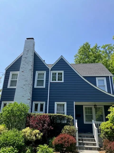This is an image from J & S Painting Plus, located in New York. The exterior photo shows a two-story home painted in a deep blue color with white trim and a tall white brick chimney.