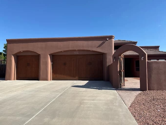 This is an image from New Mexico's Painting Specialists, located in New Mexico. The photo shows a Southwestern-style home with a smooth brown stucco exterior, three wooden garage doors, and an arched entryway under a clear blue sky.