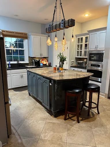 This is an image from Southern Dropcloth Painting LLC, located in Louisiana. The image shows a modern kitchen with white cabinets, a gray island featuring a marble countertop, industrial-style pendant lights, and beige tile flooring.