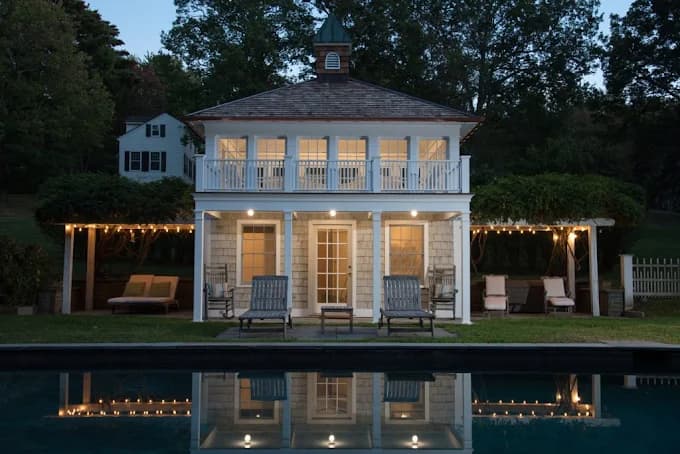 This is an image from Firefly Painting Inc, located in Colorado. The exterior photograph shows a two-story white pool house with glowing interior lights, pergola seating areas, and its reflection in the water.