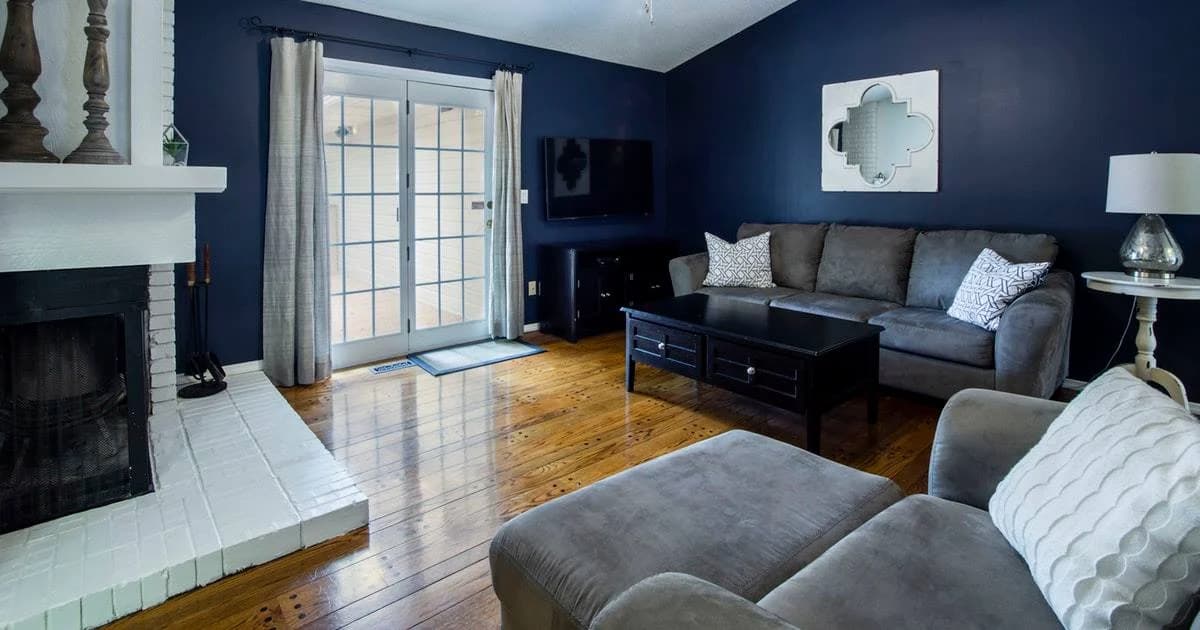 "This is an image from Texas Star Painting, located in Texas. The living room features deep navy walls, a white brick fireplace, gray furniture, and polished wood floors."