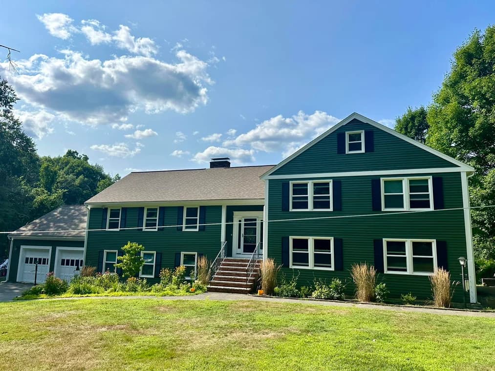 A green-painted two-story home with black shutters and a gray roof, featuring a wide lawn and landscaped garden in front.