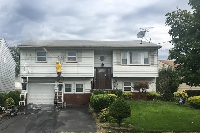 This is an image from By Hand Painting LLC, located in New Jersey. The photo shows a worker in a yellow rain jacket standing on a ladder while power washing the exterior of a two-story house with white siding and a brick foundation.