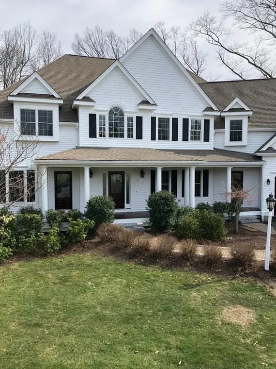 This is an image from Decolores Painting, showing a large white colonial-style house with black shutters and multiple gables. It has a covered front porch with columns and a landscaped front yard.