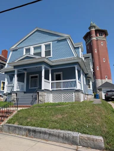 This is an image from Fresh Coat Painters of Andover, located in Massachusetts. The photo shows a freshly painted two-story blue house with white trim, a covered porch, and a nearby historic brick tower in the background.