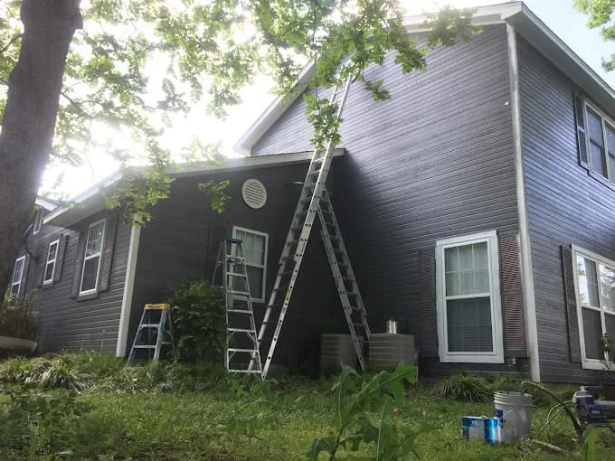 This is an image from Tyler Hoffman Painting, LLC. The picture shows a residential home with dark gray siding being painted, with tall ladders propped against the house and paint supplies at the base.