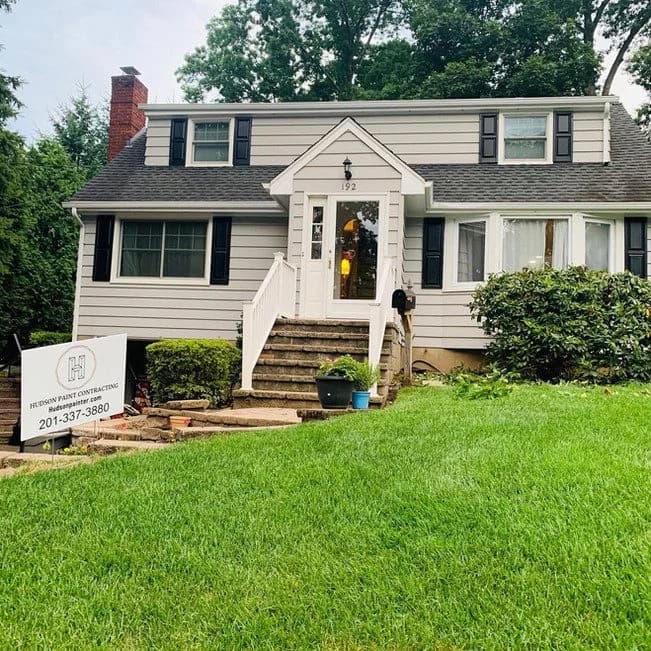 A freshly painted beige and white home exterior with black shutters, completed by Hudson Paint Contracting, featuring a yard sign with the company’s contact information.