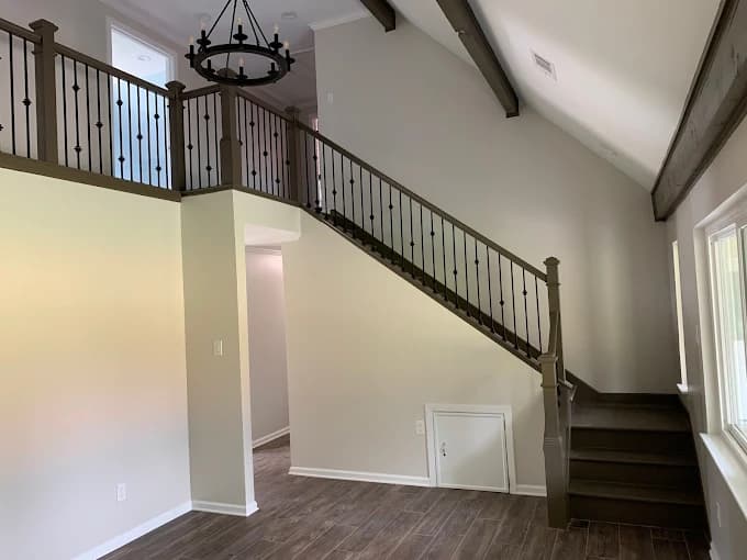 This is an image from 360 Painting of Lafayette, located in Louisiana. The photo shows a freshly painted interior staircase with dark wooden steps and railing, light beige walls, and a vaulted ceiling accented by exposed beams and a black chandelier.