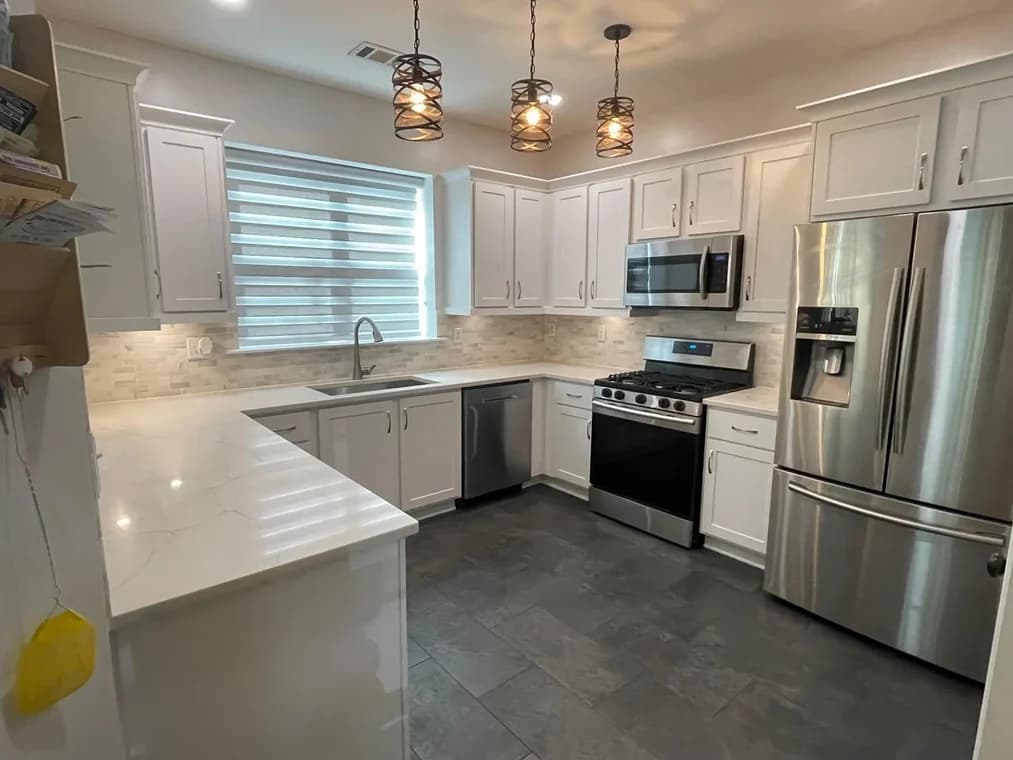 This is an image from Ortolani & Sons Painting Company, located in Massachusetts. The image shows a remodeled kitchen with freshly painted white cabinets, modern hardware, stainless steel appliances, pendant lighting, and clean neutral finishes.