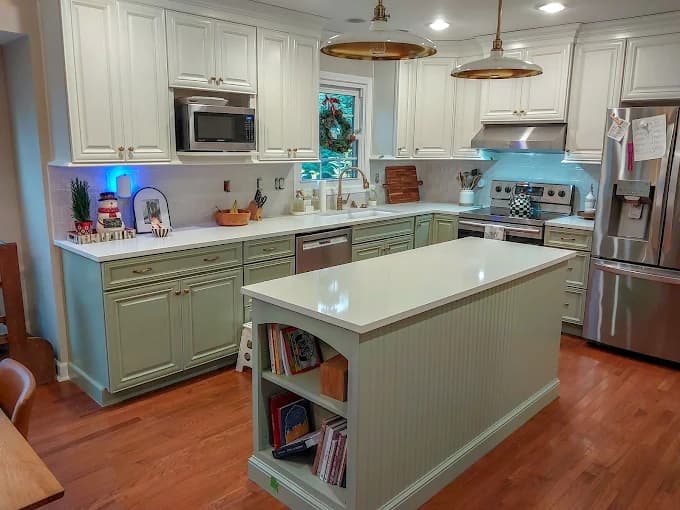 This is an image from Spray-Net Lexington, located in Kentucky. The image shows a kitchen cabinet refinishing project featuring white upper cabinets, sage green lower cabinets, and a large center island.