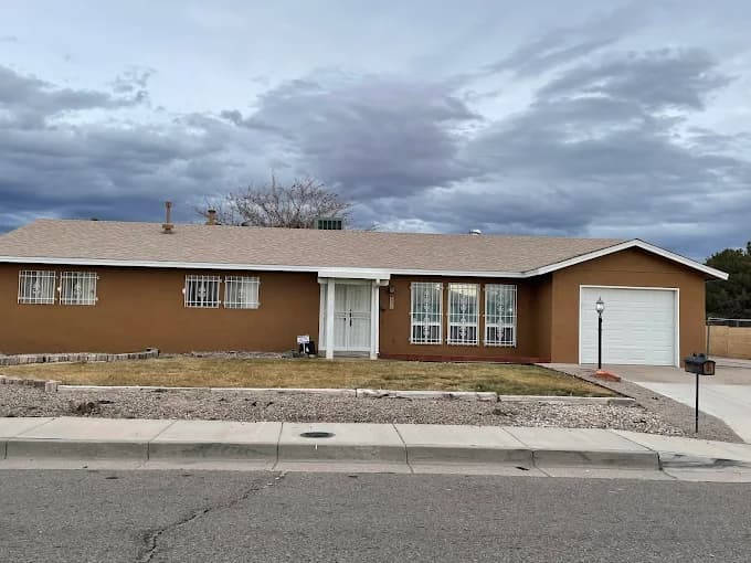 This is an image from Busby Finishes, located in New Mexico. The photo shows a single-story home with a freshly painted brown stucco exterior, white trim, and a clean desert-style front yard under an overcast sky.