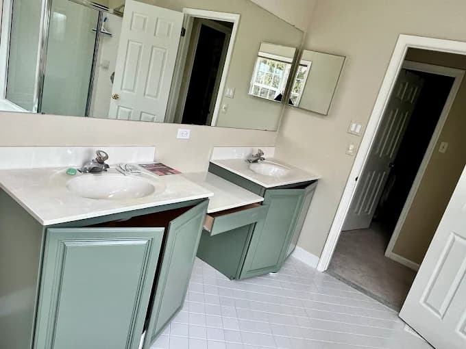This is an image from Pro Painters Plus LLC company, located in Perkasie, PA. The image is a freshly painted bathroom with sage green vanity cabinets, dual sinks, large wall mirrors, and light tile flooring in a modern home interior.