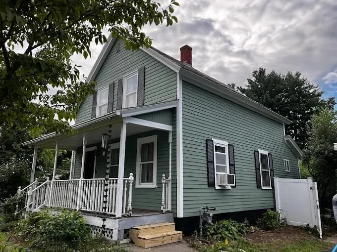 This is an image from Pancione Painting Plus LLC, located in Massachusetts. The photo shows a freshly painted light green house with white trim and black shutters, featuring a front porch and a tidy yard.