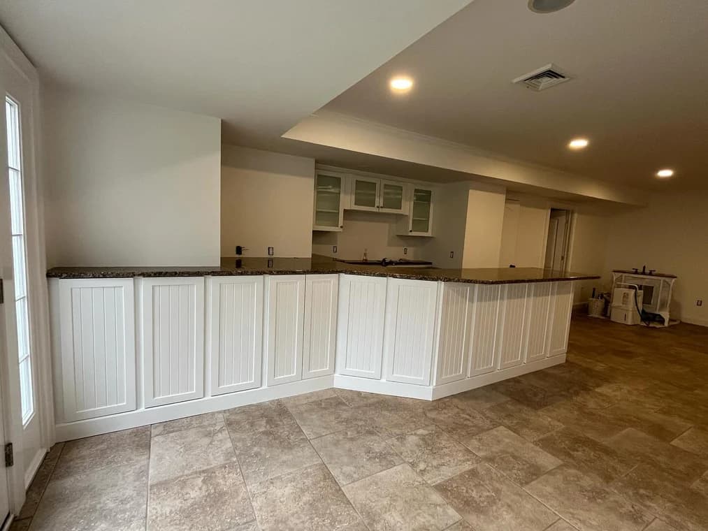 Spacious basement with tiled flooring, freshly painted white cabinetry, and a long countertop bar area.