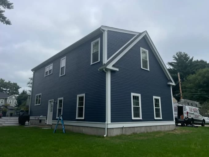 This is an image from JK Painting Service, located in Massachusetts. The photo shows a two-story home with freshly painted navy blue siding and crisp white trim, demonstrating the company’s professional exterior painting work.