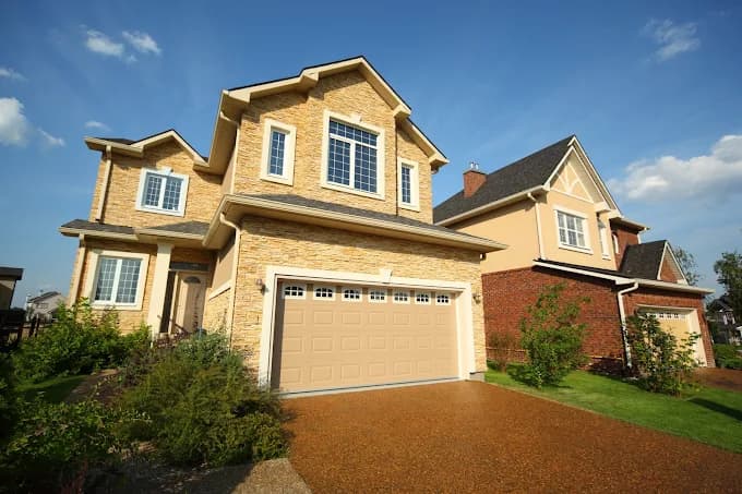 This is an image from Next Door Painting, located in Texas. The exterior photo shows a stone-front two-story home with beige trim and a clean, well-painted garage door under a clear blue sky.