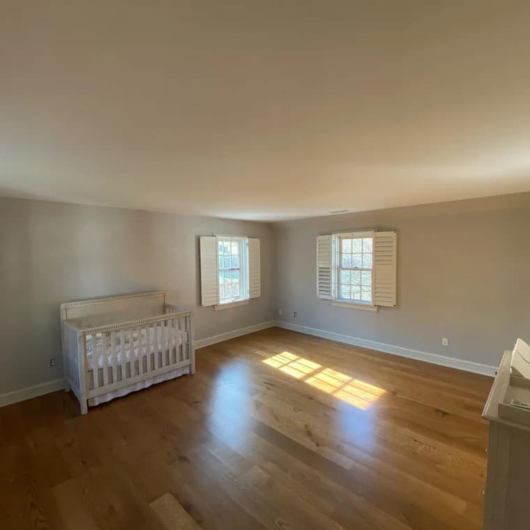 This is an image from Benner & Sons Inc company, located in Newtown Square, PA. The image is a freshly painted nursery or bedroom with light gray walls, white trim, natural wood flooring, white plantation shutters, and a white crib
