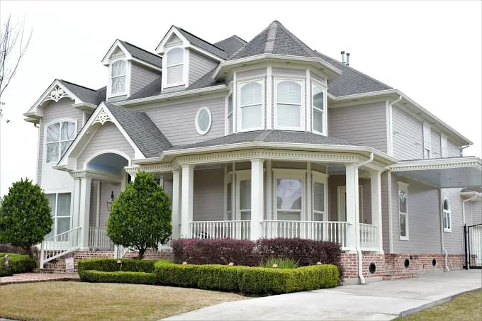 This is an image from Pelican Paint Group, located in Louisiana. The photo shows a large two-story home with light gray siding, white trim, and a wraparound porch, showcasing a clean and freshly painted exterior with manicured shrubs and a neat driveway.