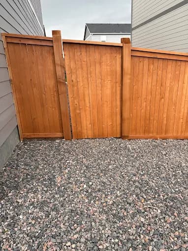 This is an image from Neighborly Fence Staining, LLC company, located in Arvada, CO The image is a newly stained vertical wooden privacy fence with varying panel heights, set between two homes with decorative gravel ground cover, completed by Neighborly Fence Staining