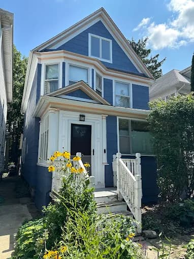 This is an image from Advantage Paint Services, located in New York. The image shows the exterior of a blue painted two-story home with white trim, a covered front porch, and landscaped front yard.