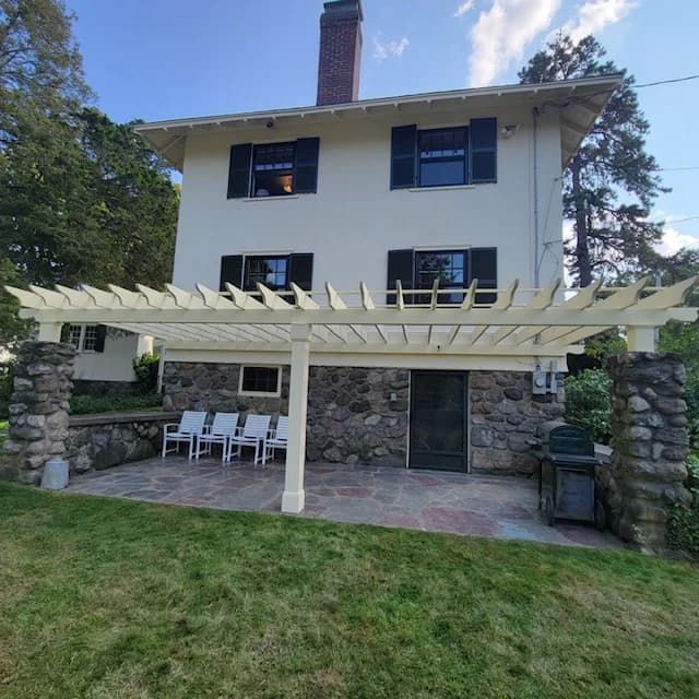 This is an image from CertaPro Painters of North Shore and Cape Ann, located in Massachusetts. It captures a three-story home with light cream siding, dark shutters, and a backyard pergola painted in a clean white finish.