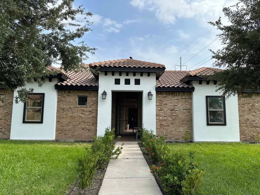 "This is an image from Armored Painting, located in Texas. The house exterior features painted white stucco sections, light brick walls, dark trim, and a walkway framed by green landscaping."
